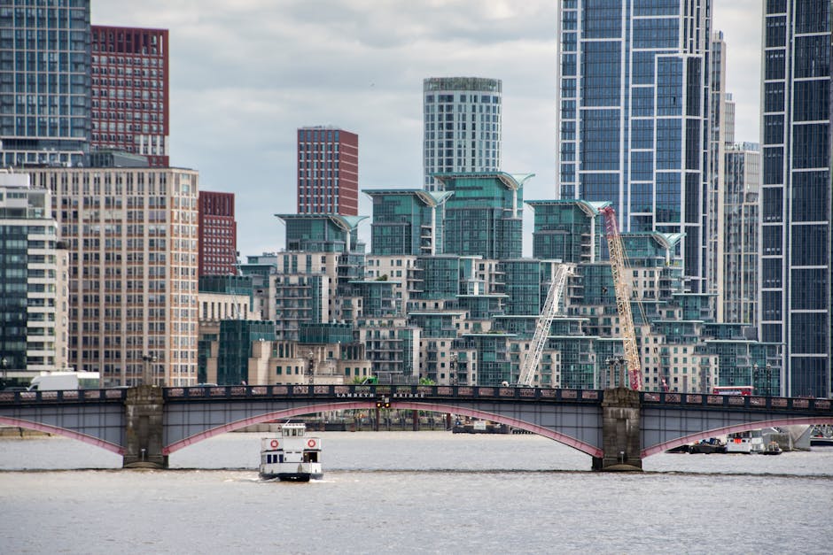A cityscape view showing modern office buildings with glass facades and high-rise structures against a partly cloudy sky. In the foreground, there's an elevated railway track with an orange and grey p